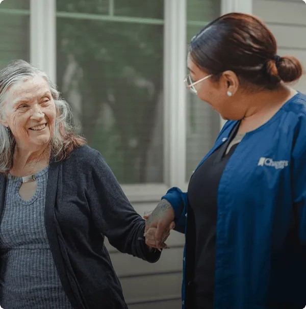An older woman smiles while holding hands with a caregiver in a blue uniform, standing outdoors in front of a building with large windows.