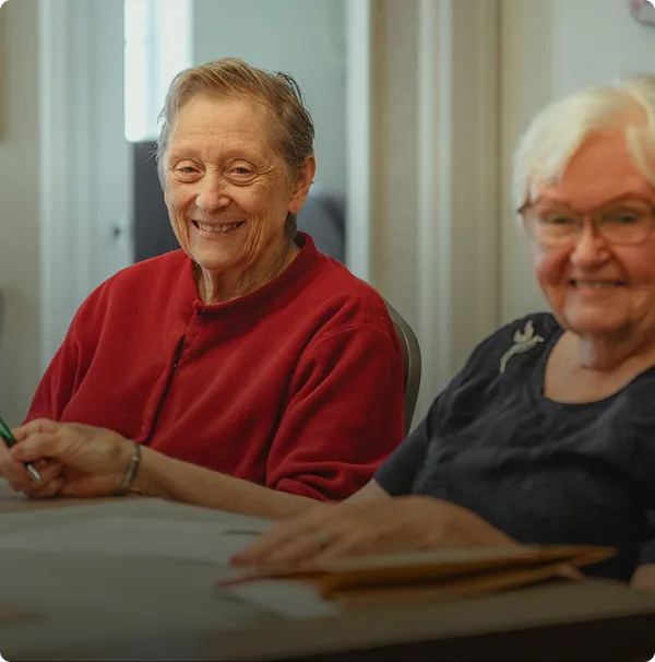 Two elderly women sit at a table, smiling and holding pens, appearing to enjoy a shared activity in a well-lit room.